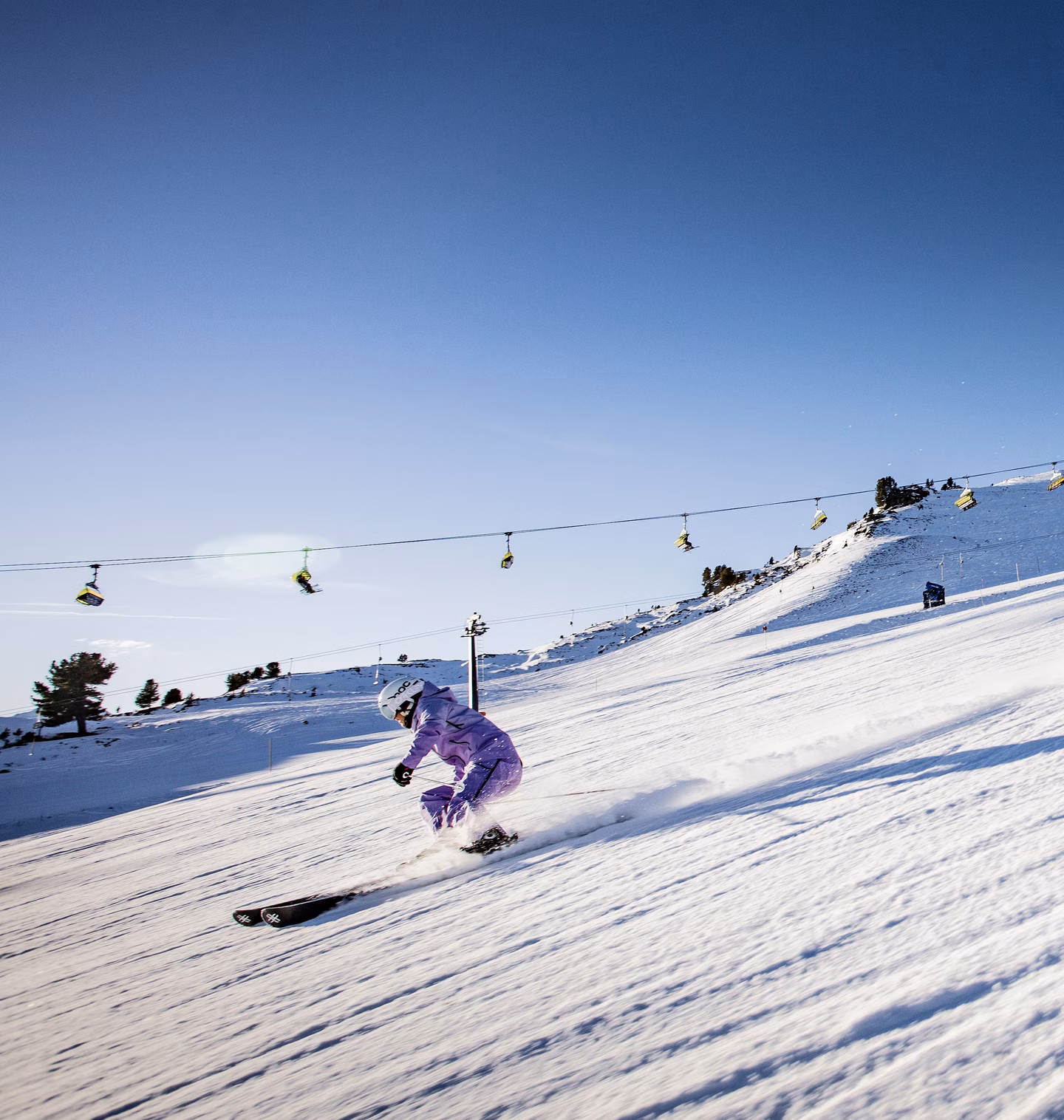 Zwei Skifahrer carven dicht hintereinander eine sonnige Piste hinunter, im Hintergrund ein Sessellift über verschneite Hänge.