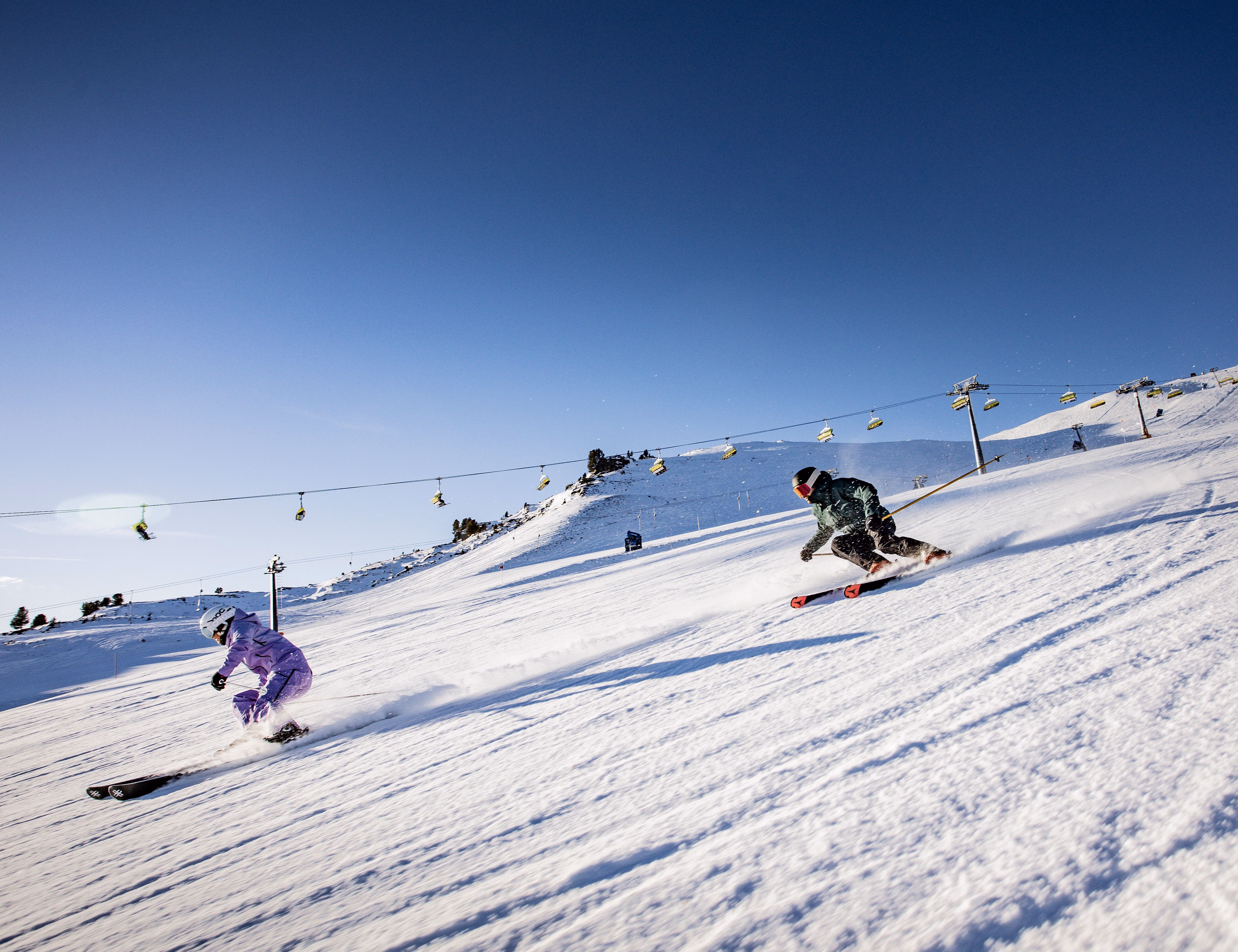 Zwei Skifahrer carven dicht hintereinander eine sonnige Piste hinunter, im Hintergrund ein Sessellift über verschneite Hänge.