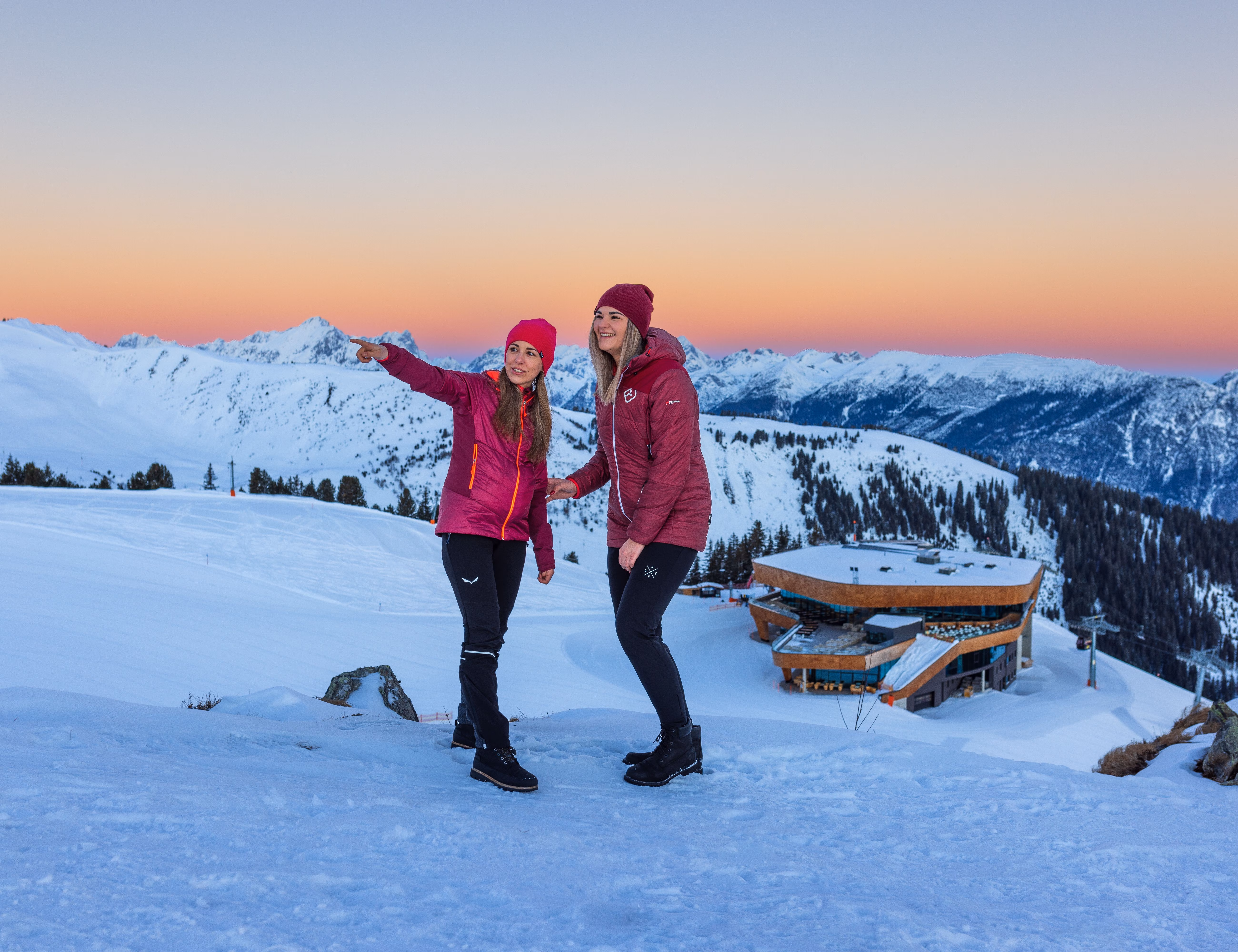 Zwei Frauen in Winterjacken stehen bei Dämmerung auf dem verschneiten Spieljoch mit Bergpanorama und Bergstation im Zillertal